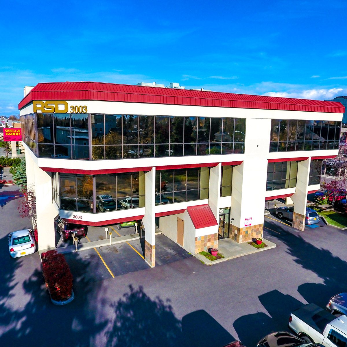 Exterior of RSD Properties' commercial office space at 3003 Minnesota Drive on a sunny Anchorage, Alaska day. White three-story building with red accents and large tinted windows across the second and third floors. Parking area in front and side with several cars. The RSD logo with "3003" on the upper left of the building. Buildings and a Wells Fargo sign are in the background left side.