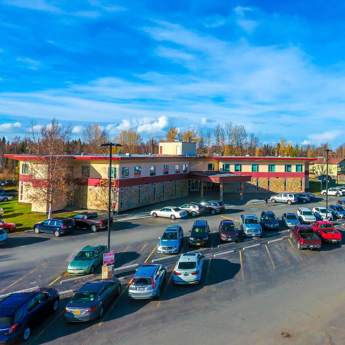Exterior of RSD Properties' commercial office space at 4050 Lake Otis Parkway on a sunny Anchorage, Alaska day. Tan two-story building with red accents and small office windows across the top. Parking area in front with several cars. A lawn is to the left of the building and trees are in the background.