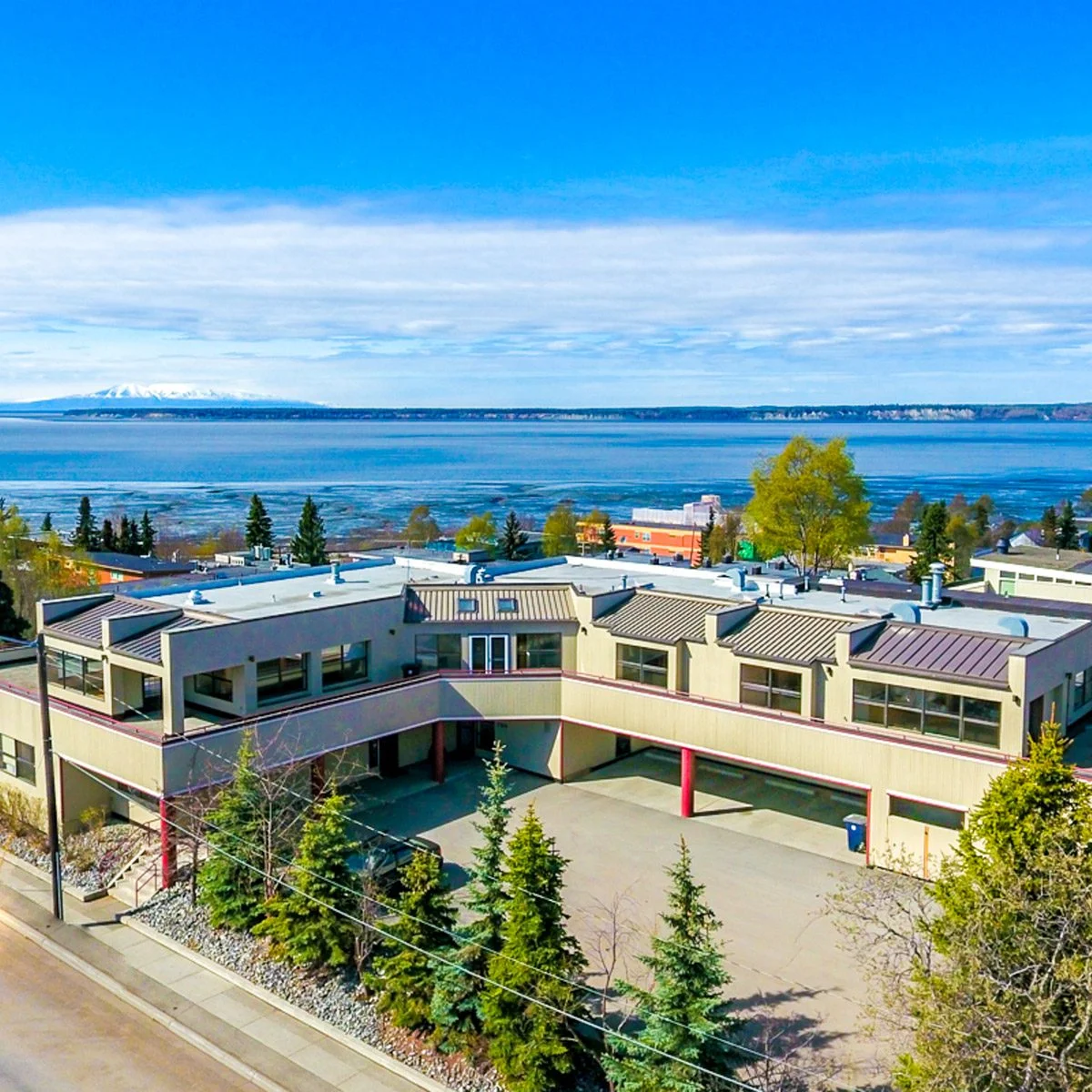 Exterior of RSD Properties' commercial office space at 731 N Street on a sunny Anchorage, Alaska day. Tan two-story L-shaped building with trees lining the parking area in the front and a view of Cook Inlet in the background.