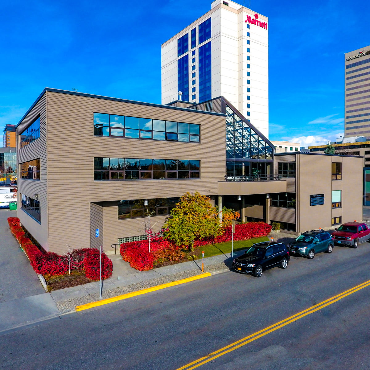 Exterior of RSD Properties' coworking space, Cowork by RSD, on a sunny Anchorage, Alaska day. Brown building with an atrium in the middle and red and green hedges around the left and front sides. Cars are parked on street in front, and the Marriot and Conoco Phillips building are in the background
